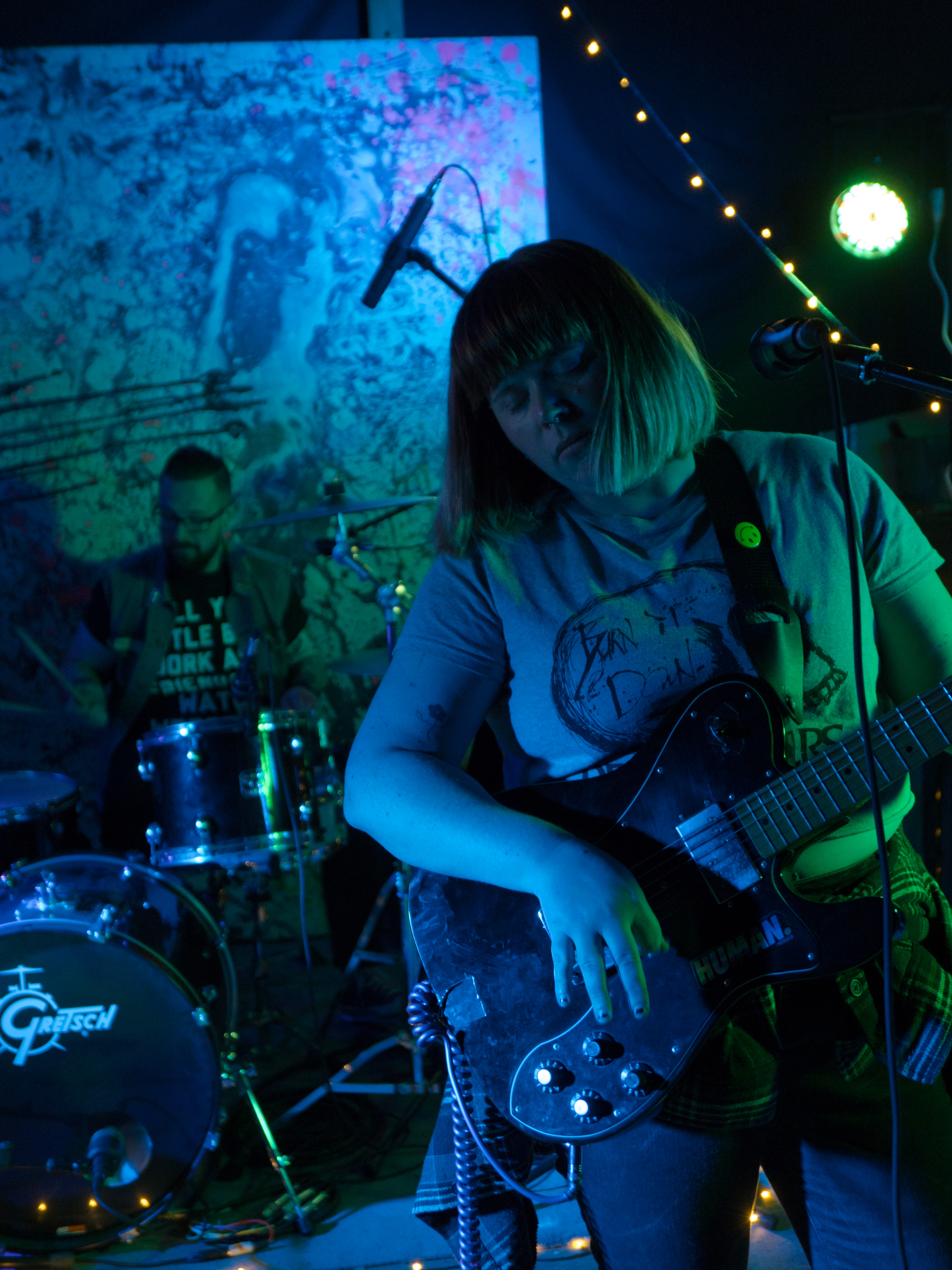 Bridget stands in the front of the stage, holding her guitar. She has a red-blonde bob with a blunt fringe & she looks lost in the music. Andy is visible at the drums behind her.